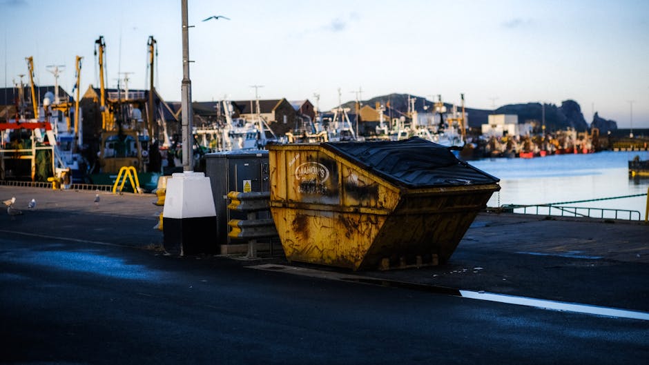A large, yellow metal skip with visible rust and black plastic lid sits on a paved area near a marina, surrounded by utility boxes and electrical equipment. The skip is positioned next to a white and