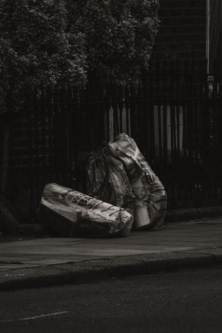 A large pile of mixed waste consists of various discarded items including cardboard boxes, plastic bags, paper, and packaged goods, all overflowing from a grey recycling bin marked for mixed paper and